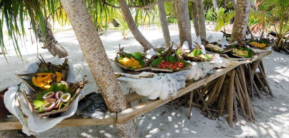 Tropical outdoor setting with wooden table holding plates of fresh fruit and large seashells under palm trees on sandy ground.