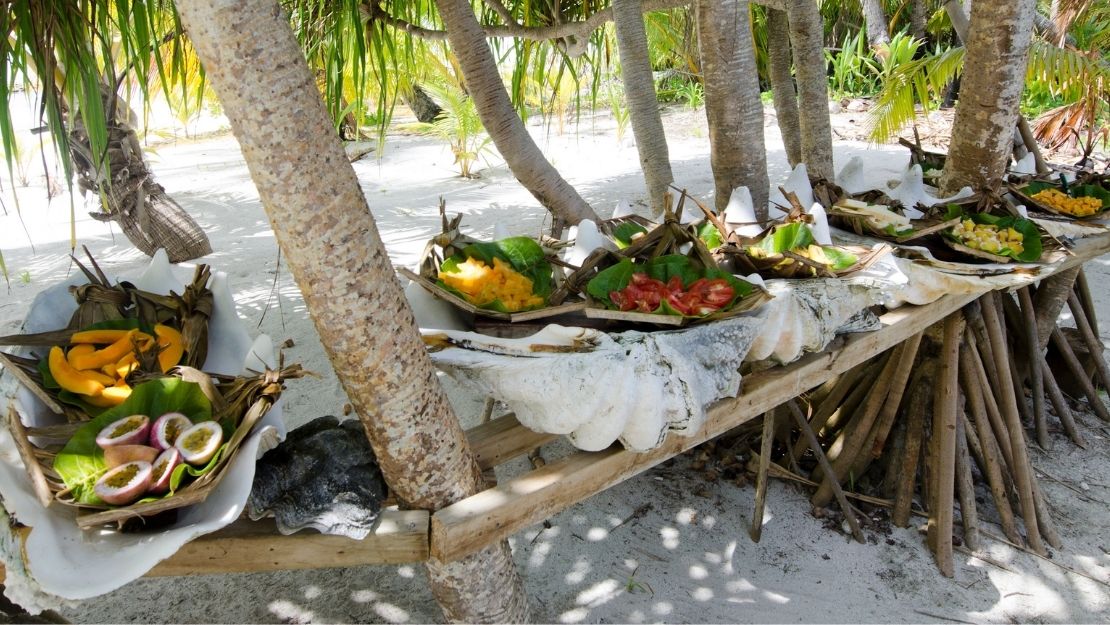 Tropical outdoor setting with wooden table holding plates of fresh fruit and large seashells under palm trees on sandy ground.