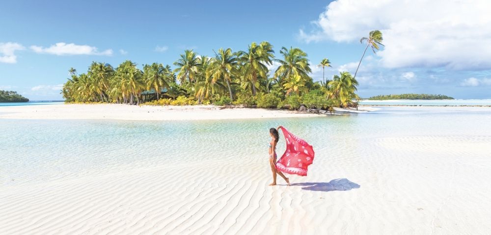 Woman walking on a white sandy beach near clear shallow water with palm trees under a bright blue sky.