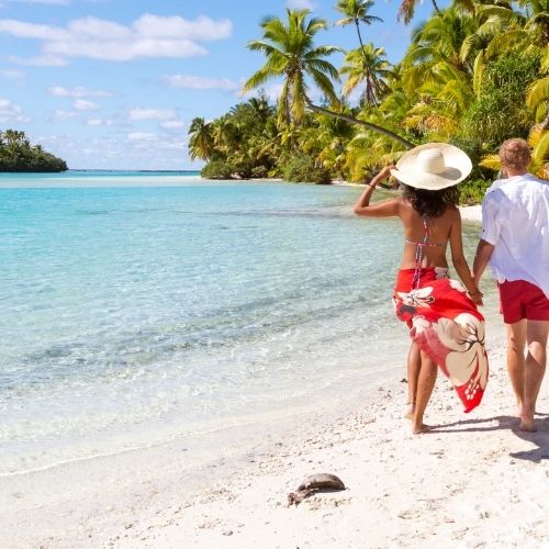 Couple walking hand in hand along a tropical beach with clear blue water and palm trees.