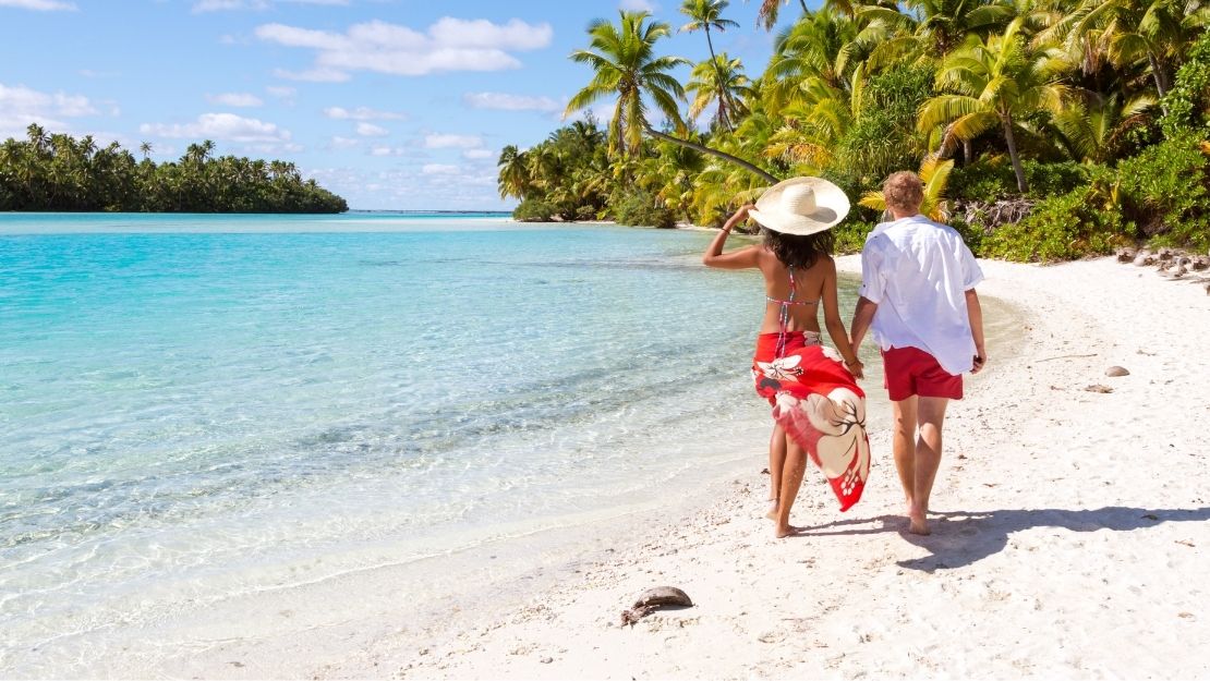 Couple walking hand in hand along a tropical beach with clear blue water and palm trees.