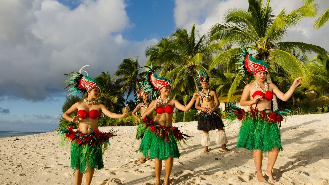 Traditional Polynesian dancers in colorful costumes perform on a sandy beach with palm trees under a partly cloudy sky.