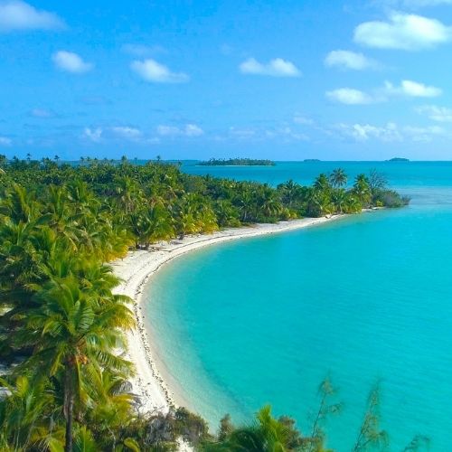 A picturesque tropical beach with clear turquoise water, sandy shores, and lush palm trees under a blue sky with scattered clouds.