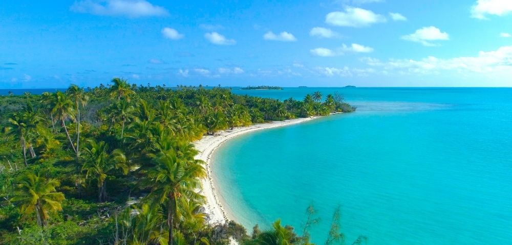 A picturesque tropical beach with clear turquoise water, sandy shores, and lush palm trees under a blue sky with scattered clouds.