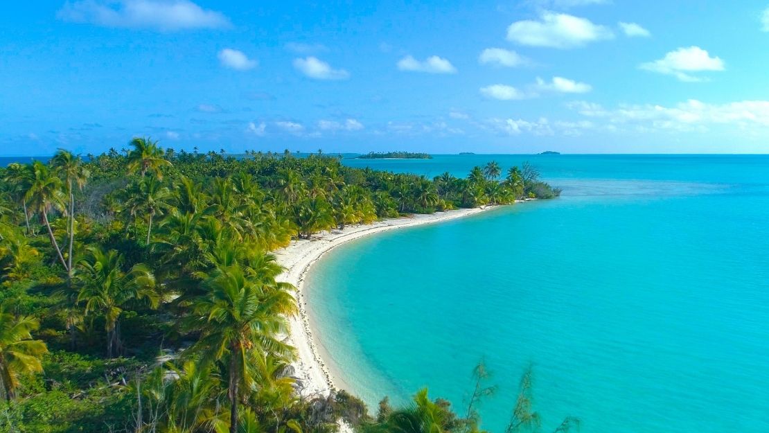 A picturesque tropical beach with clear turquoise water, sandy shores, and lush palm trees under a blue sky with scattered clouds.