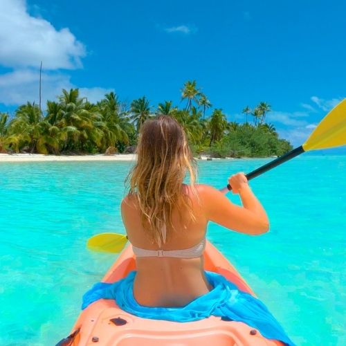 A person kayaking in clear turquoise waters near a tropical island, surrounded by palm trees.