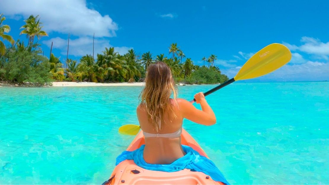 A person kayaking in clear turquoise waters near a tropical island, surrounded by palm trees.