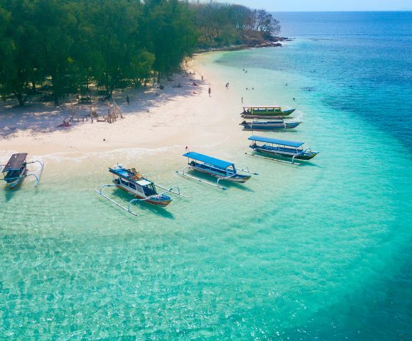 Aerial view of a tropical beach with clear turquoise water, white sand, and lush green trees, dotted with several outrigger boats and a few people swimming in the shallow sea.