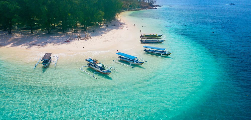 Aerial view of a tropical beach with clear turquoise water, white sand, and lush green trees, dotted with several outrigger boats and a few people swimming in the shallow sea.