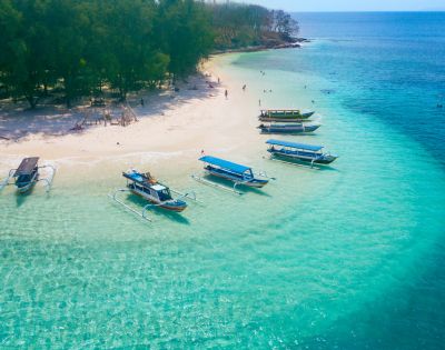 Aerial view of a tropical beach with clear turquoise water, white sand, and lush green trees, dotted with several outrigger boats and a few people swimming in the shallow sea.