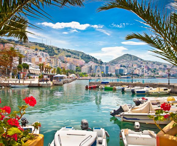 A vibrant harbor with numerous boats moored in clear turquoise water, a coastal city with colorful buildings cascading down a hill under a bright blue sky with fluffy clouds, and lush palm fronds framing the foreground.