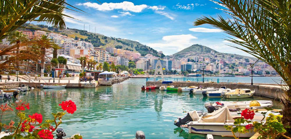 A vibrant harbor with numerous boats moored in clear turquoise water, a coastal city with colorful buildings cascading down a hill under a bright blue sky with fluffy clouds, and lush palm fronds framing the foreground.