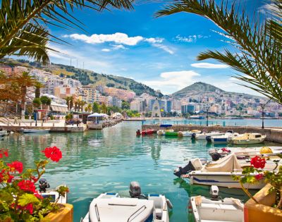 A vibrant harbor with numerous boats moored in clear turquoise water, a coastal city with colorful buildings cascading down a hill under a bright blue sky with fluffy clouds, and lush palm fronds framing the foreground.