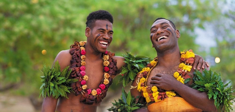 Two men wearing traditional floral garlands and greenery, laughing joyfully outdoors with a blurred green background.