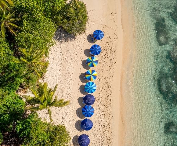 Aerial view of a sandy beach with colorful umbrellas arranged in a line near the water's edge, surrounded by greenery.