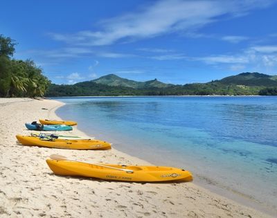 Kayaks on a sandy beach by calm waters with lush greenery and hills in the background.