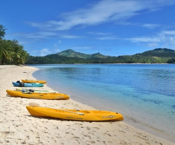 Kayaks on a sandy beach by calm waters with lush greenery and hills in the background.
