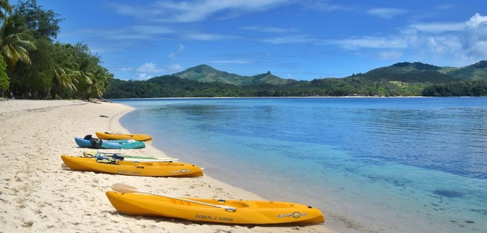 Kayaks on a sandy beach by calm waters with lush greenery and hills in the background.
