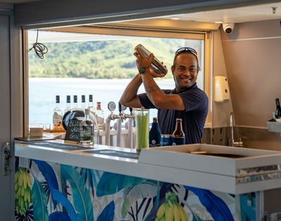 A bartender shaking a cocktail at a bar with bottles and views of the water in the background.