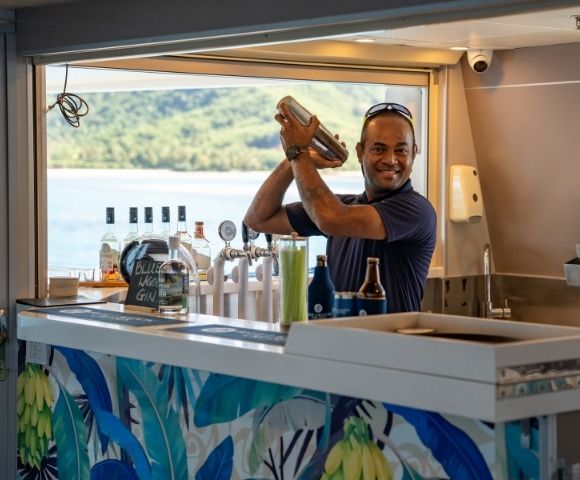 A bartender shaking a cocktail at a bar with bottles and views of the water in the background.