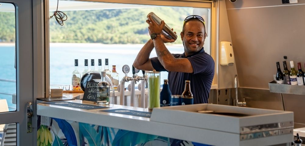 A bartender shaking a cocktail at a bar with bottles and views of the water in the background.