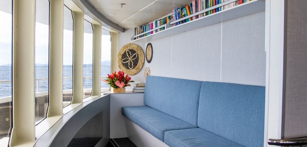 A cozy seating area with a blue couch, bookshelves filled with books, and decorative items by large windows overlooking the ocean.
