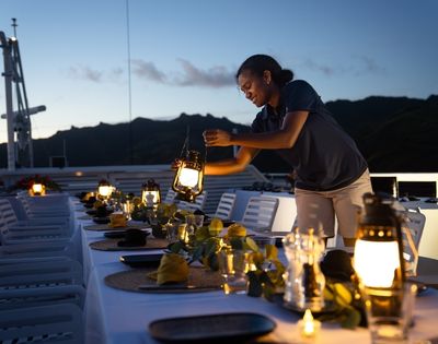 A woman setting up a dining table with lanterns and decorations on a boat during sunset.