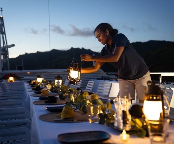 A woman setting up a dining table with lanterns and decorations on a boat during sunset.