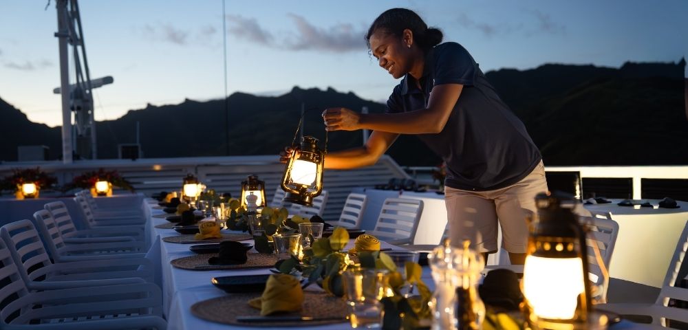 A woman setting up a dining table with lanterns and decorations on a boat during sunset.