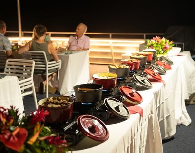 A buffet setup with various dishes served in red pots on a table, surrounded by guests dining in the background.