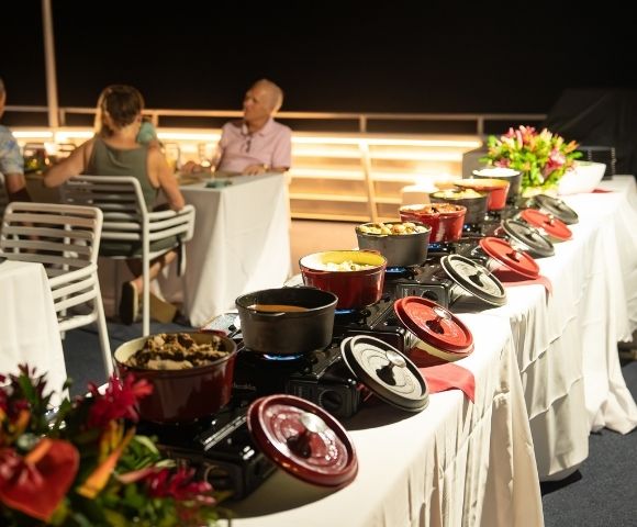 A buffet setup with various dishes served in red pots on a table, surrounded by guests dining in the background.