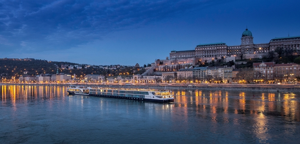 River cruise ship sailing on calm water with illuminated historic buildings and a hill under a blue evening sky.