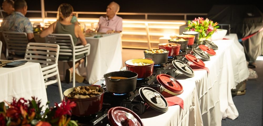 A buffet setup with various dishes served in red pots on a table, surrounded by guests dining in the background.