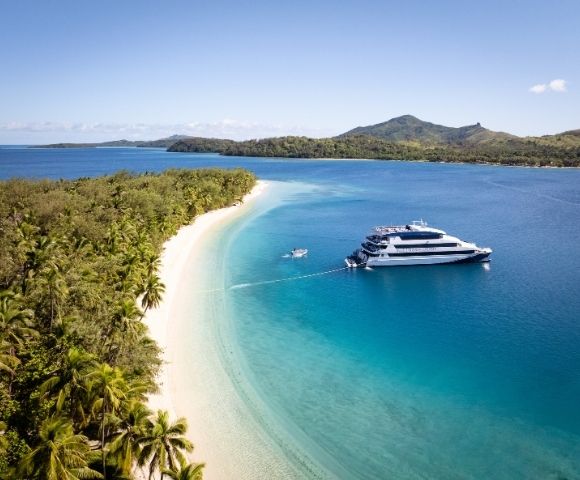 A luxury yacht anchored near a tropical shore with a sandy beach and lush greenery, surrounded by clear blue water and mountains in the background.