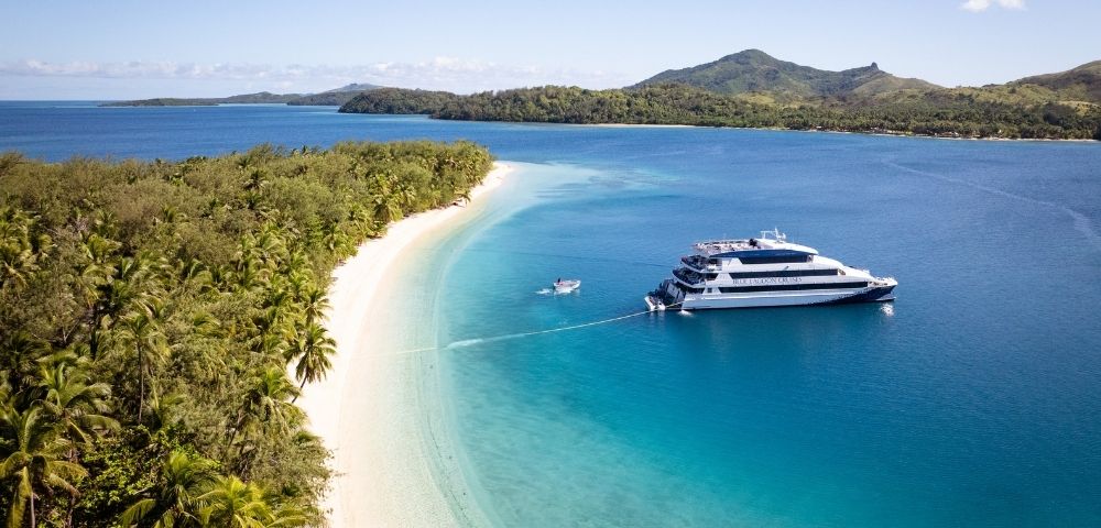 A luxury yacht anchored near a tropical shore with a sandy beach and lush greenery, surrounded by clear blue water and mountains in the background.