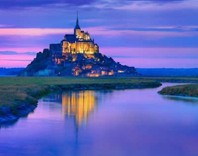 A picturesque view of Mont Saint-Michel at sunset, with vibrant purple and orange hues in the sky, reflecting in the tranquil waters of the surrounding area.
