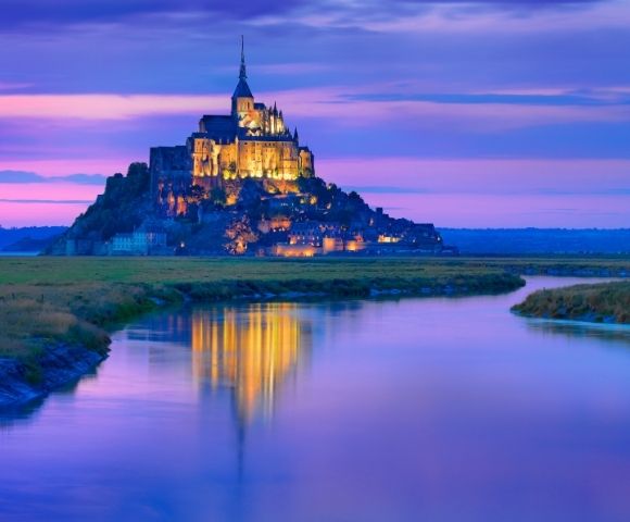 A picturesque view of Mont Saint-Michel at sunset, with vibrant purple and orange hues in the sky, reflecting in the tranquil waters of the surrounding area.
