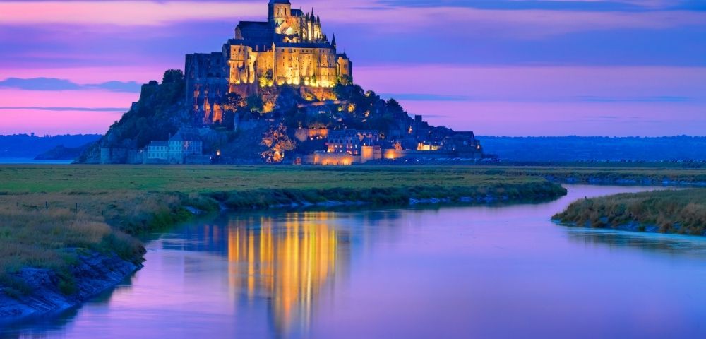 A picturesque view of Mont Saint-Michel at sunset, with vibrant purple and orange hues in the sky, reflecting in the tranquil waters of the surrounding area.