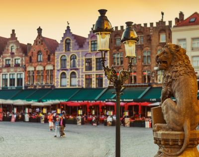 Stone lion statue in a European square with historic buildings and outdoor cafes with red umbrellas in the background during sunset.