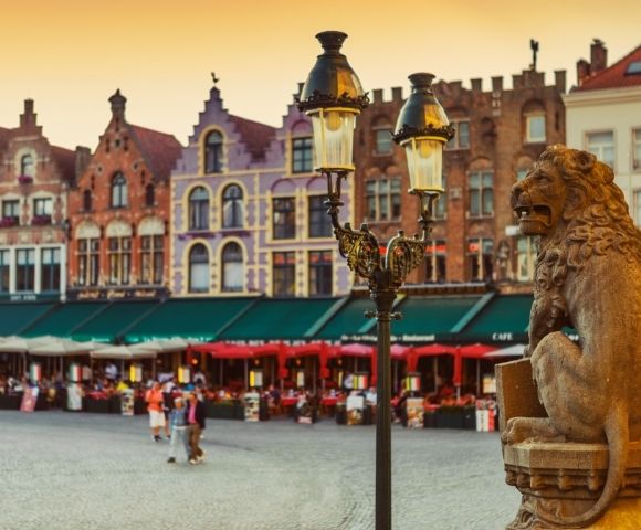 Stone lion statue in a European square with historic buildings and outdoor cafes with red umbrellas in the background during sunset.