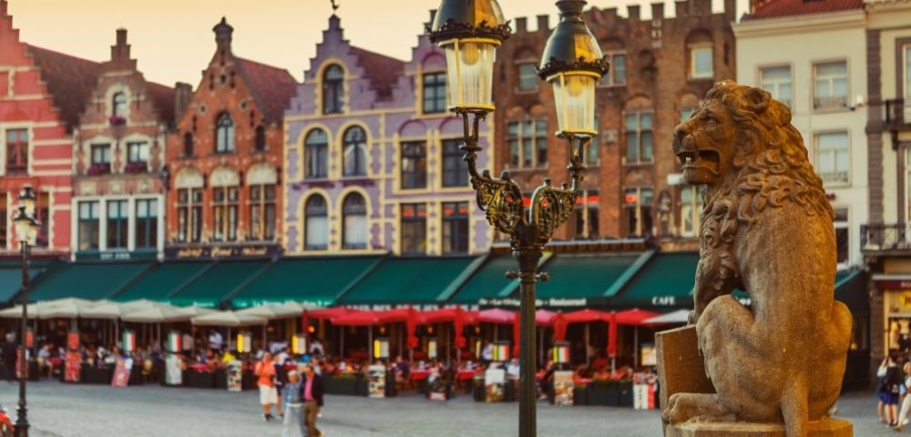 Stone lion statue in a European square with historic buildings and outdoor cafes with red umbrellas in the background during sunset.