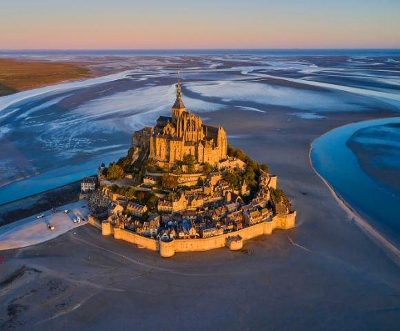 An aerial view of a medieval abbey and village perched on a rocky island surrounded by tidal flats and shimmering blue water under a warm sunset sky.