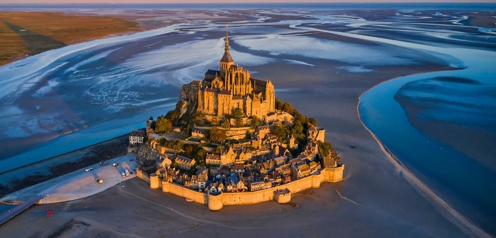 An aerial view of a medieval abbey and village perched on a rocky island surrounded by tidal flats and shimmering blue water under a warm sunset sky.