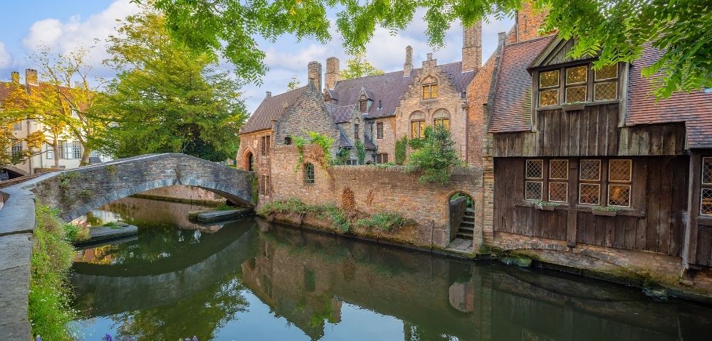 A stone bridge arches over a tranquil canal reflecting historic brick buildings and trees.