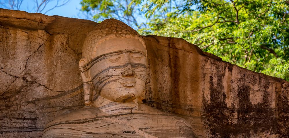 A large, ancient stone carving of a reclining Buddha statue, showing the face and upper body, carved into a rock face with layered striations, under a canopy of green trees and blue sky.
