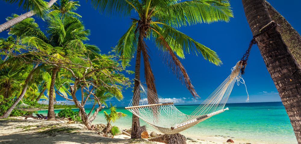 A white rope hammock hangs between two palm trees on a sandy tropical beach, overlooking clear turquoise water under a bright blue sky.
