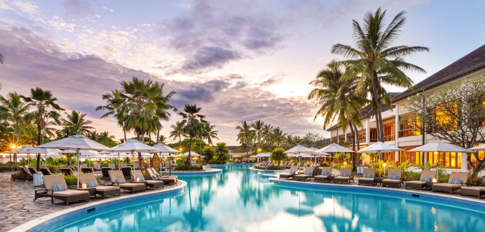 A wide, luxurious resort swimming pool with turquoise water, surrounded by rows of lounge chairs and umbrellas, with lush palm trees and a building in the background under a soft, cloud-filled sunset sky.