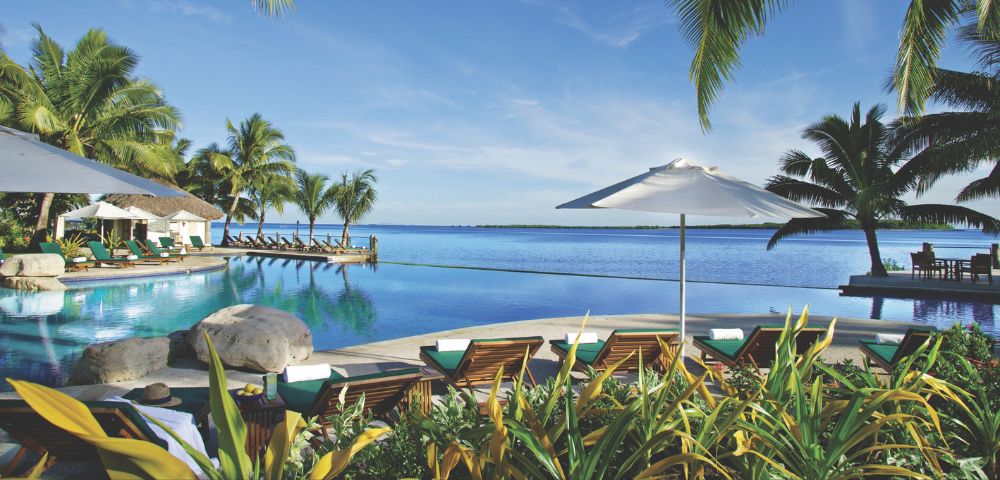 Tropical resort with an infinity pool overlooking a calm blue ocean, surrounded by palm trees, lounge chairs, and umbrellas.