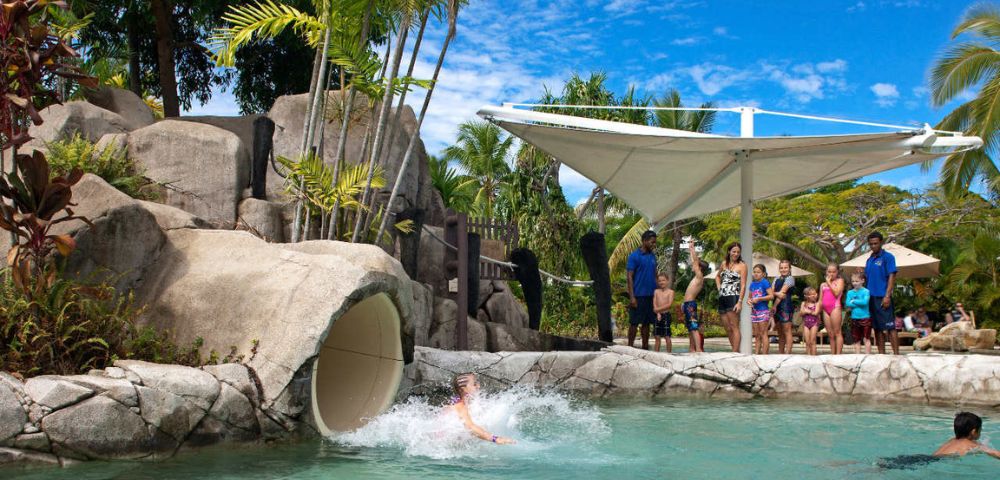 A person splashes into a swimming pool as they exit a water slide built into artificial rock formations, with palm trees and a large white shade canopy overhead, and a group of people standing near the slide entrance.
