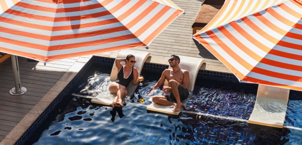 Two people relax in shallow in-pool loungers under orange and white striped umbrellas at a sunny resort.